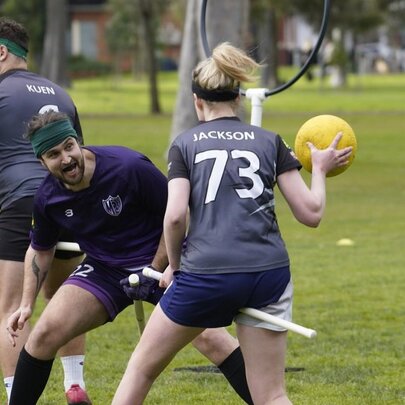 Three people are playing a game of quadball on a grassy field, one of them is holding the yellow ball.