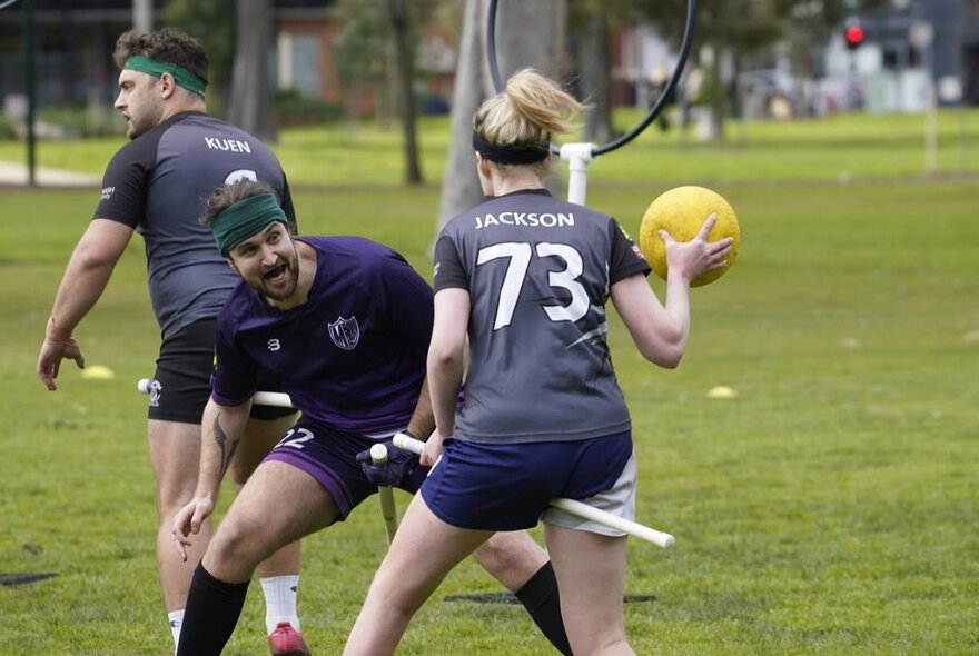 Three people are playing a game of quadball on a grassy field, one of them is holding the yellow ball.