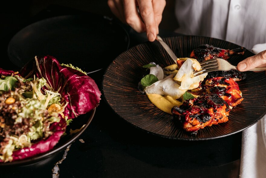 Hands holding a knife and fork above a plate of food.