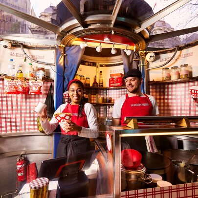 Two staff members wearing red aprons inside La Petite Creperie, with red and white checked decor and serving counter.