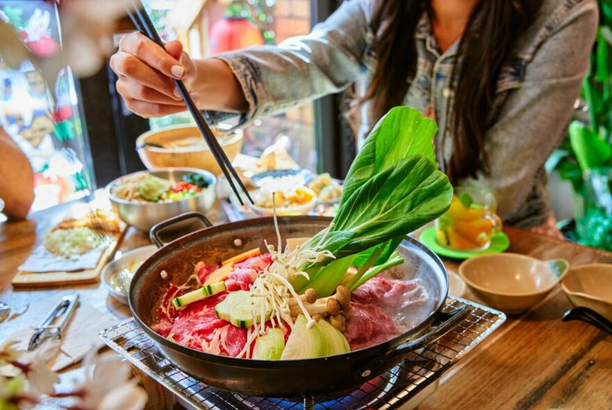 A dining table laden with dishes, including a pot on a small burner, with a diner's hand leaning  in to add fresh ingredients to it.