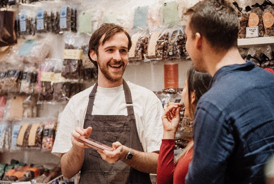 A person in a shop showing two customers a product against a wall lined with food products.