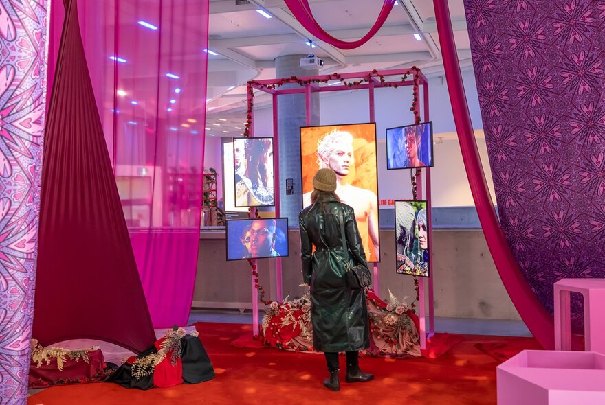 A woman in a leather trench coat looking at screens showing a man, surrounded by pink curtains.
