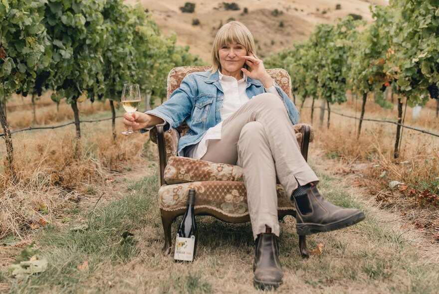 A woman sitting in an armchair in the middle of a vineyard, a glass of wine in her hand and a bottle on the ground. 