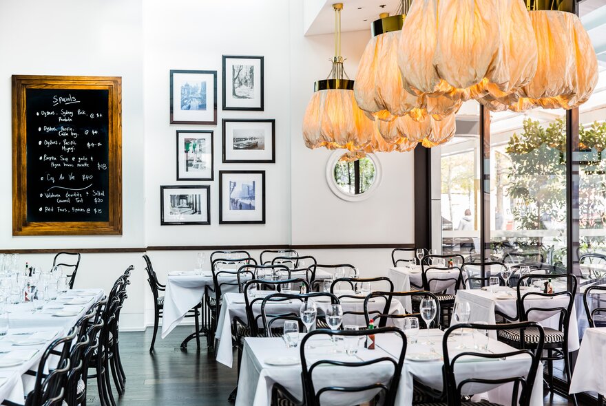 Bright and airy restaurant interior with white walls, white tablecloths, black bistro-style chairs, a chalkboard menu on the wall and pendant light fixtures hanging from the ceiling. 