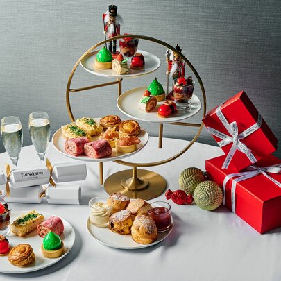 An array of Christmas-themed High Tea sweet and savoury treats on a white table alongside champagne glasses and two red-wrapped presents.
