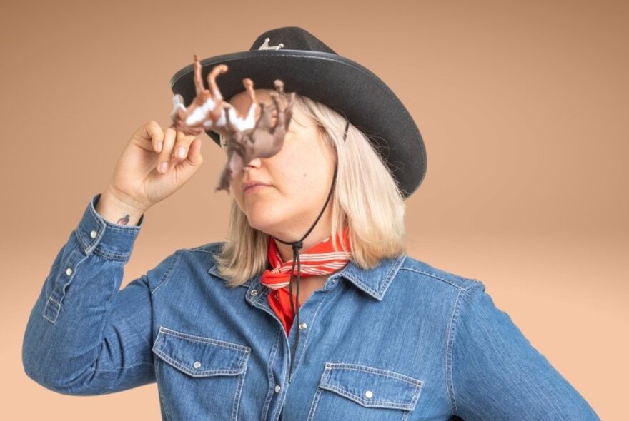 Performer wearing a denim shirt and cowboy hat, holding plastic horses to her eyes.