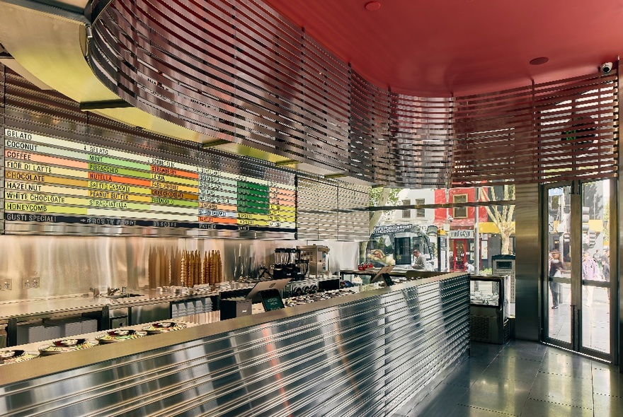 Interior of Piccolina gelato store with a curved stainless steel serving counter, reflected in a curved stainless steel lighting design suspended from the ceiling.