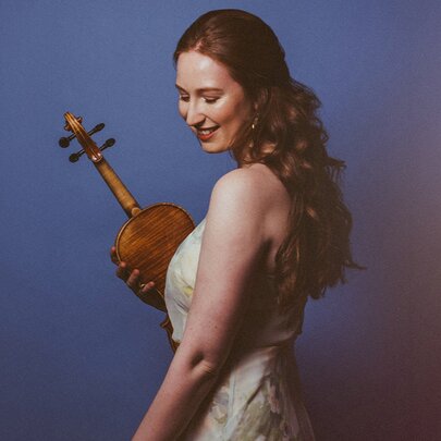 Violinist Augusta McKay Lodge looking over her shoulder holding her violin in her right hand. 