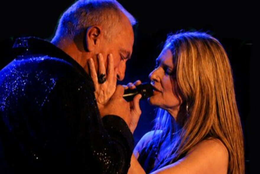 A male and female singer caressing each other's faces as they sing together on stage.