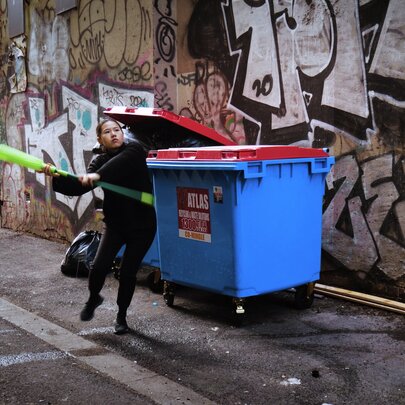 A video still from artist, Huiyi Xiao, showing a woman swinging a mop in front of a large blue wheelie-bin in a graffitied laneway in Melbourne.