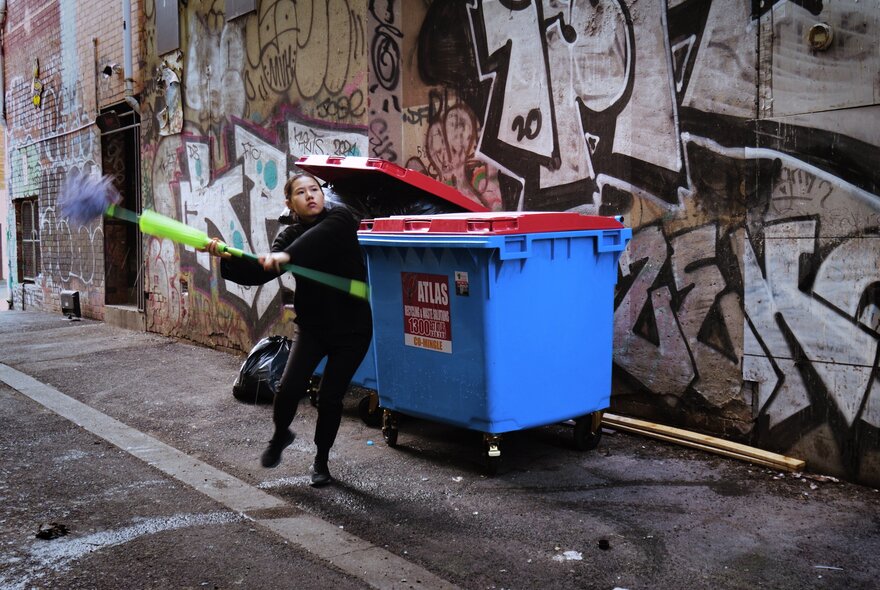 A video still from artist, Huiyi Xiao, showing a woman swinging a mop in front of a large blue wheelie-bin in a graffitied laneway in Melbourne.