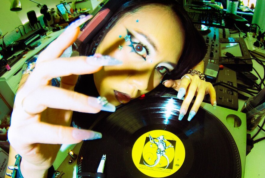 Looking down from above at a person's face with heavy eye makeup who holds a vinyl record in their hands, their fingers have long fingernails painted blue and adorned with jewels; in a music studio setting.