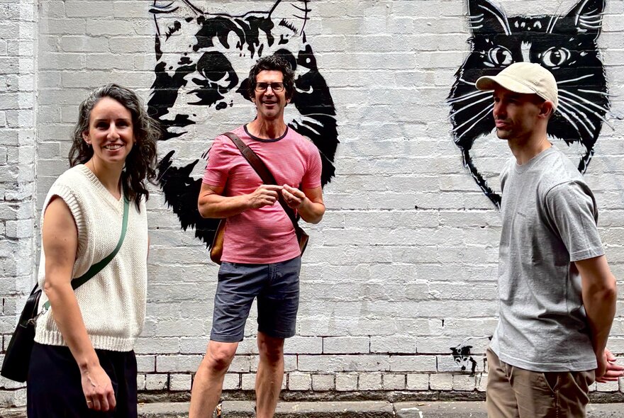 A tour guide wearing a pink t-shirt, talking to a couple on a walking tour about the mural art on the wall behind him.