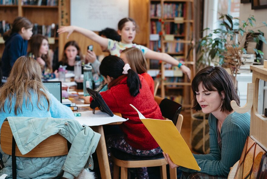 Teacher and children working at large table with pen and paper and laptops.