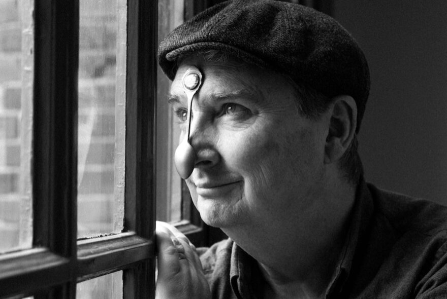 Man peering through a window, wearing a cap with a spoon resting on his nose.