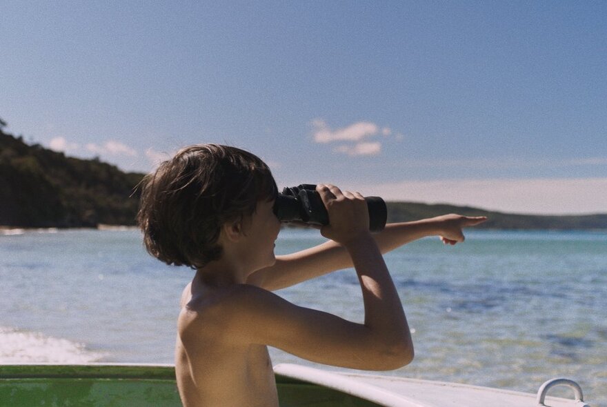 Film still of a young boy at a beach looking through binoculars and pointing to the horizon.