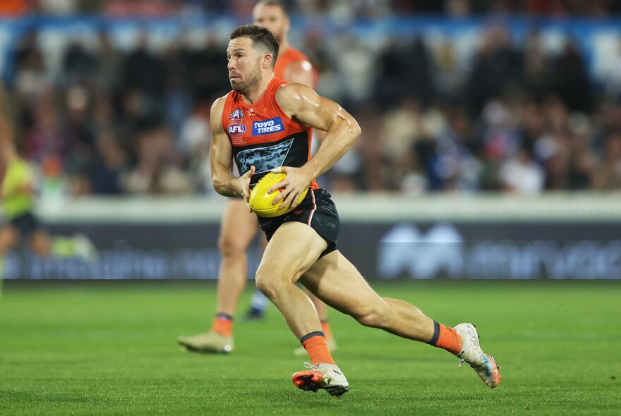 A GWS Giants AFL football player running with the ball during a match, the blurred stadium crowd in the background.
