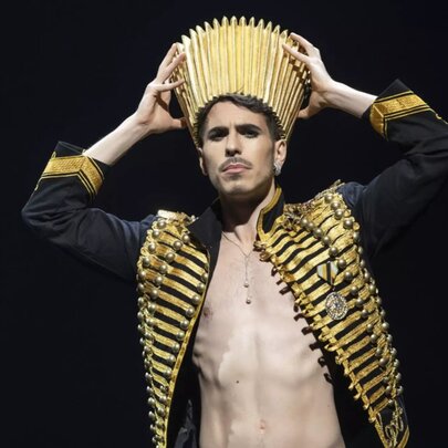 Man with bare chest exposed beneath gilded military jacket, holding a gold crown to his head.
