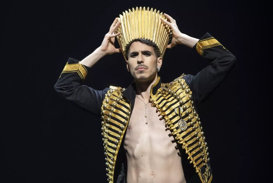 Man with bare chest exposed beneath gilded military jacket, holding a gold crown to his head.