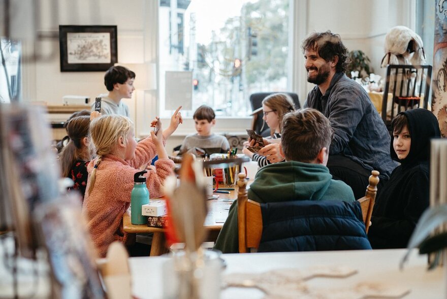 Primary school-aged children sitting around large worktables, writing and interacting with each other and their teacher in a large airy room, with windows looking out onto the street.