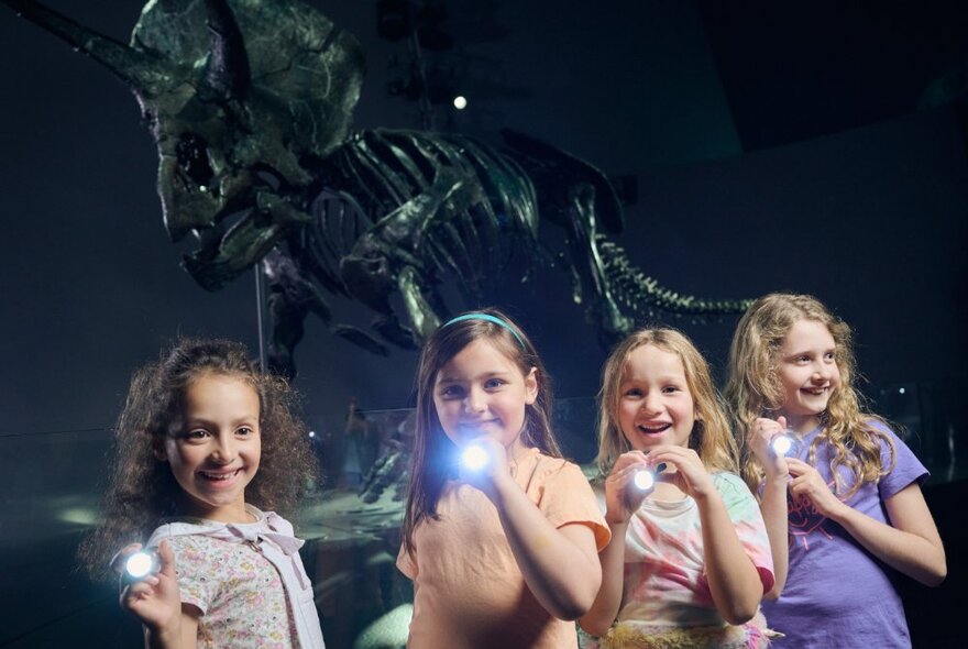 Four little girls smiling and holding torches, a dinosaur skeleton visible in the background. 