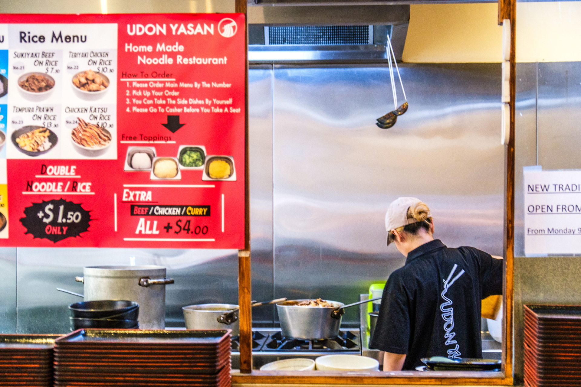 A person is behind the counter at a restaurant kitchen making udon noodles. There is a menu board behind them