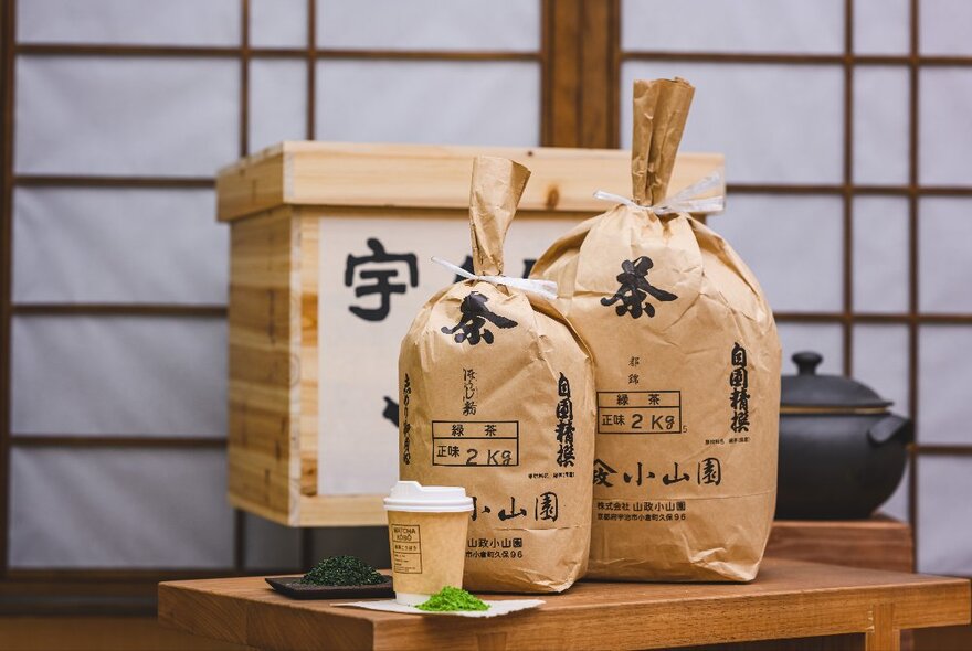 Two Japanese paper bags with Japanese characters printed on them, resting on a wooden table with Japanese paper screens behind them.
