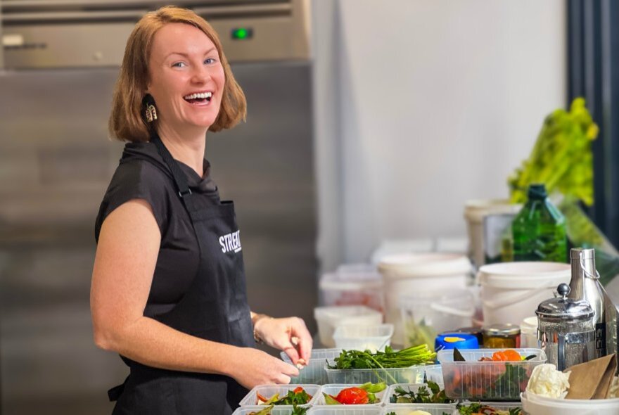 A woman in a black apron laughing as she prepares food at a bench top. 