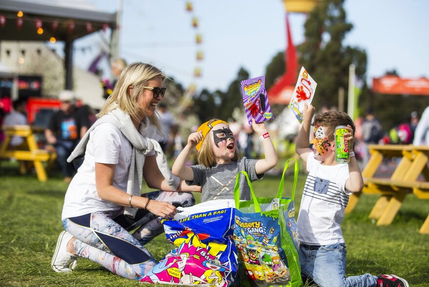 A mother with her two young children sitting on grass at the Melbourne Royal Show surrounded by showbags, carnival rides in the background.