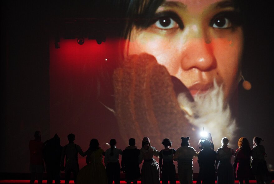 A dramatic close-up of a person's face reflecting on a screen, with the silhouette of a line of people in the foreground.