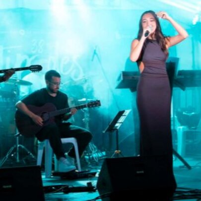 A female singer in a long dark dress performs on a lit stage with a live band playing instruments.