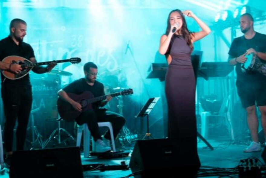A female singer in a long dark dress performs on a lit stage with a live band playing instruments.