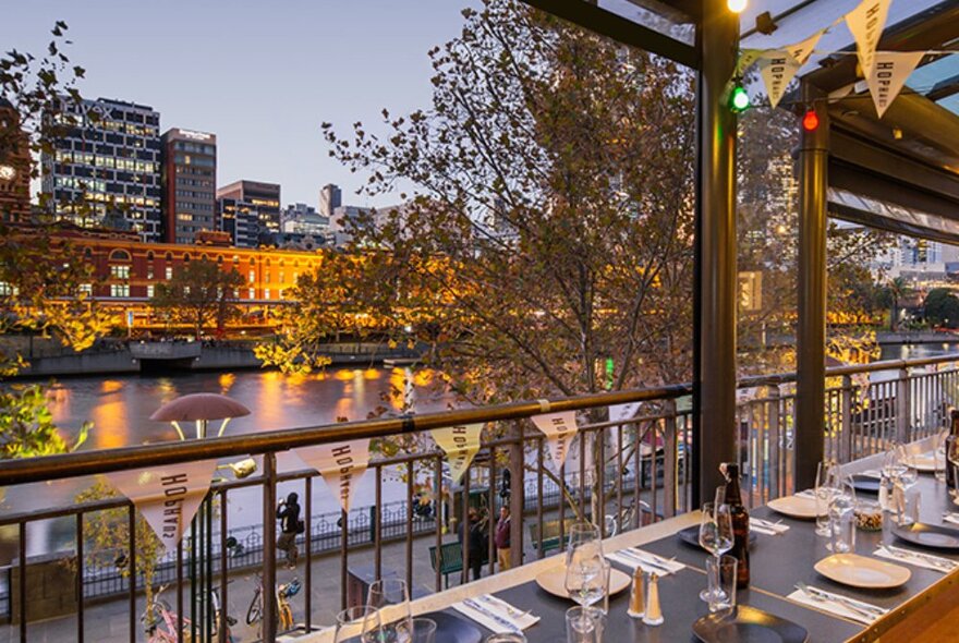 A view of Melbourne's Yarra River from the terrace of a restaurant on Southbank, trees in the foreground and Flinders St Station in the background.