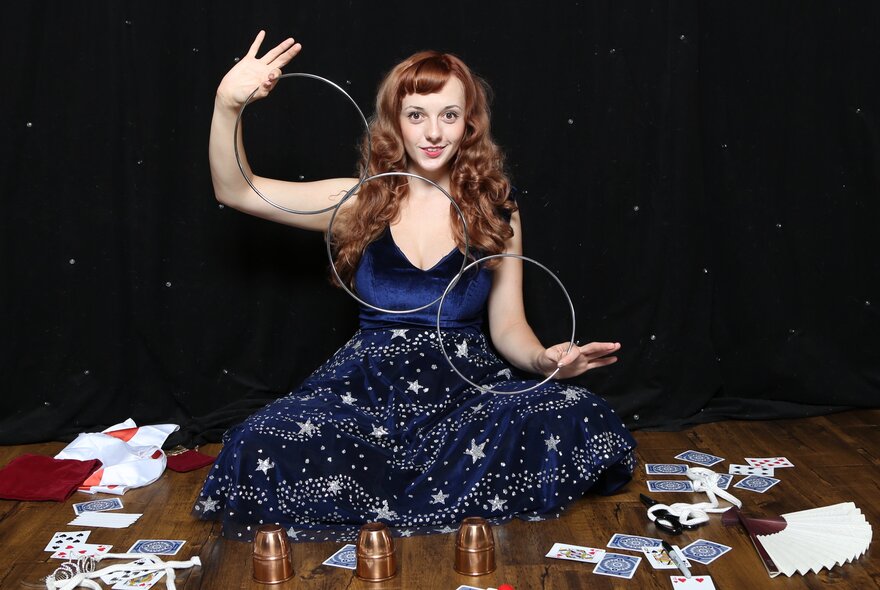 Performer Magical Katrina, seated on the ground wearing a blue dress surrounded by decks of playing cards and other magic props. 