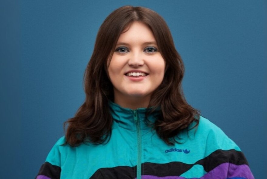 Comedian and musician Sez, smiling at the viewer, with straight brown shoulder length hair, wearing an aqua and purple zipped-up tracksuit top.