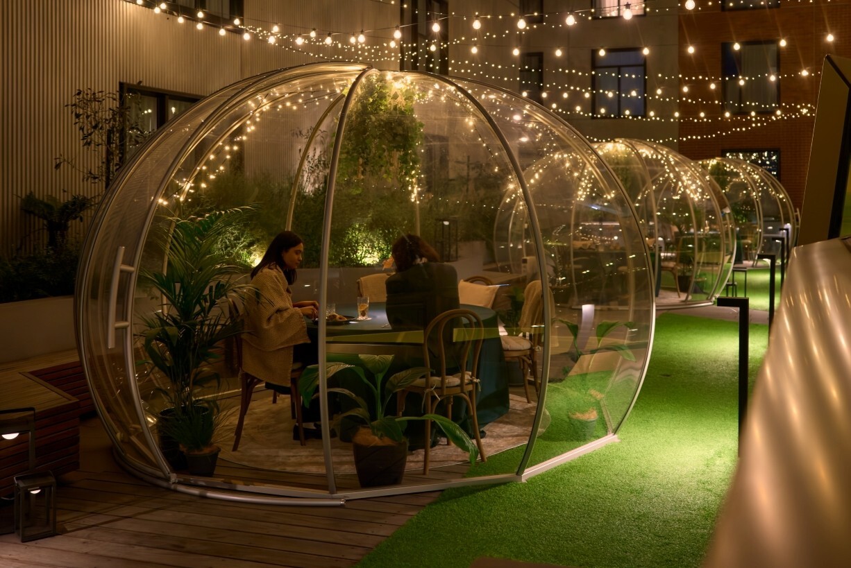 A couple sit in a see through globe in a courtyard decorated with fairylights