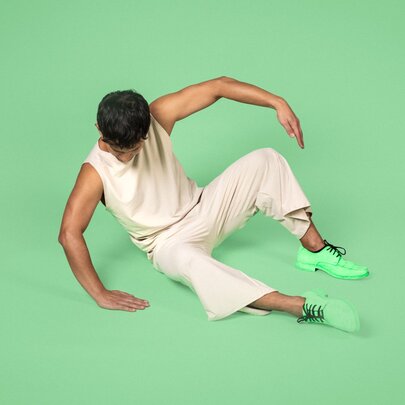 A person demonstrating a dynamic dance move while wearing a comfortable, neutral-toned sleeveless top and trousers, set against a green background.
