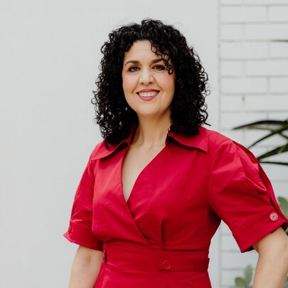 Dr Tania Cammarano, a middle-aged woman posing for the camera in a red dress against a white brick wall.