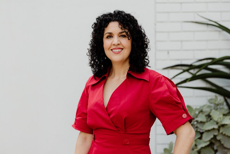 Dr Tania Cammarano, a middle-aged woman posing for the camera in a red dress against a white brick wall.