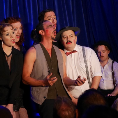 Cast members dressed in cabaret-style costumes and posing as a group on stage in  front of a blue velvet curtain.