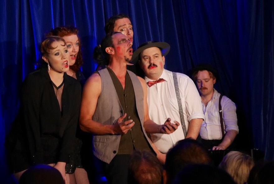 Cast members dressed in cabaret-style costumes and posing as a group on stage in  front of a blue velvet curtain.