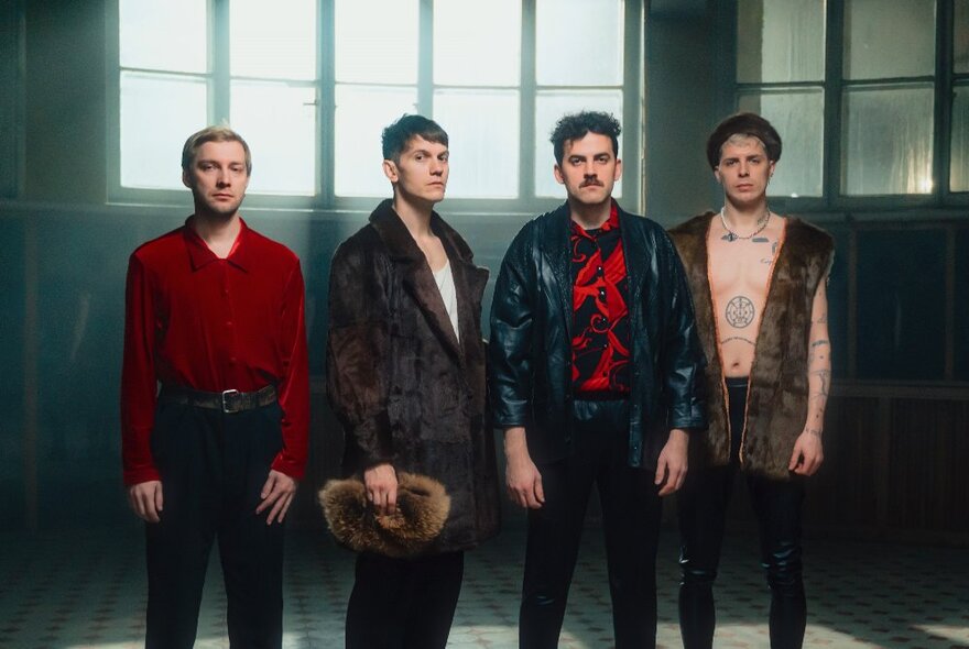 Four-member pop group standing in a sparse room with a row of windows, wearing red and black clothing.