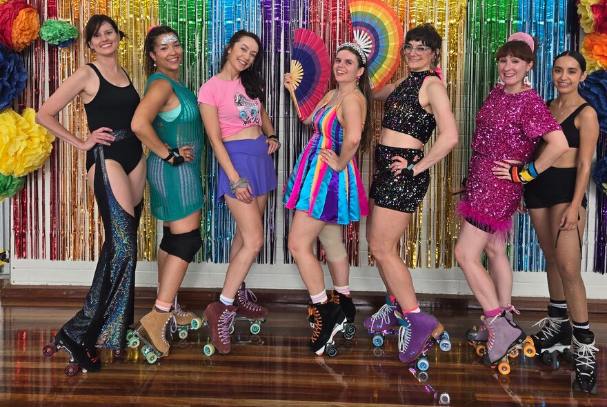 Seven colourfully dressed roller skaters posing in front of a glittery rainbow wall hanging.