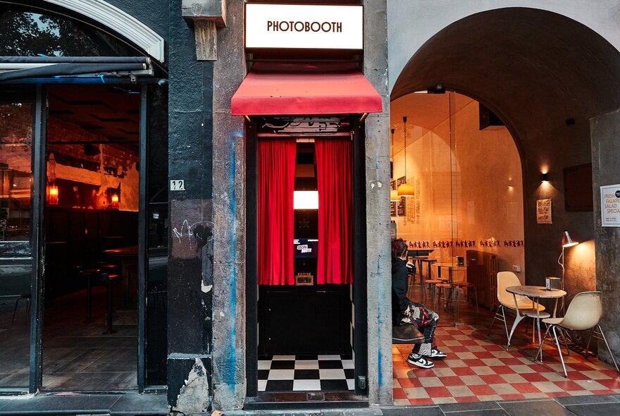 Vintage-style photo booth with red curtains and check floor, next to restaurant interior with similar design and arched windows.