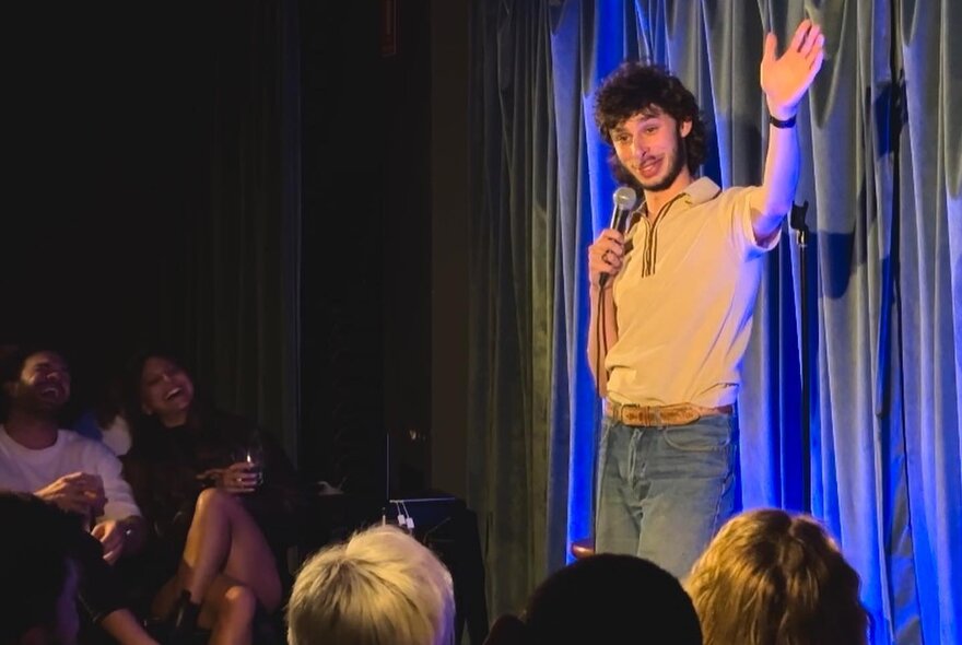 Comedian and mime artist Con Coutis performing stand-up on a stage in front of a live audience.