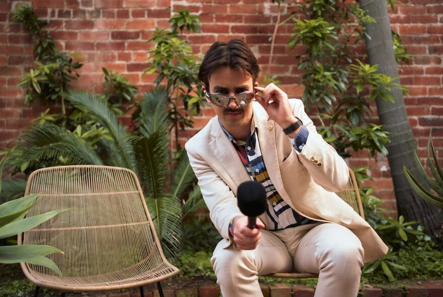 Comedian Sweeney Preston wearing a white suit and seated in a chair in an outdoor courtyard garden while holding a reporter's microphone in his hand.