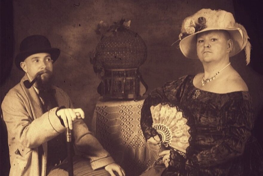 A sepia toned repro picture of a man with a hat and pipe and woman with a large hat and splayed fan. 