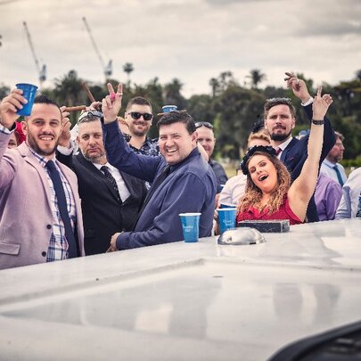 A group of racegoers on board a boat, all looking at the camera with hands raised.