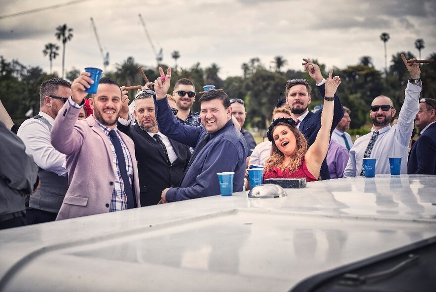 A group of racegoers on board a boat, all looking at the camera with hands raised.